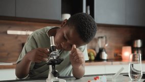 African American boy is sitting at table in kitchen, studying and doing homework. He uses microscope, discovering knowledges during homeschool. Black skinned schoolboy is interested and curious - Powered by Shutterstock - Get 15% off with code: PIKWIZARD15