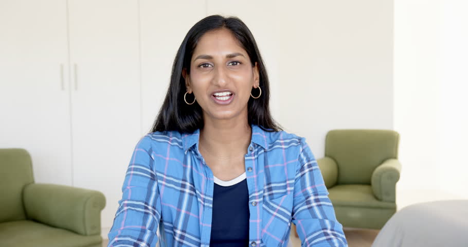Close up of people talking to the camera, smiling woman in blue plaid shirt sitting in living room, looking at camera. the person is fully visible, sitting, half body