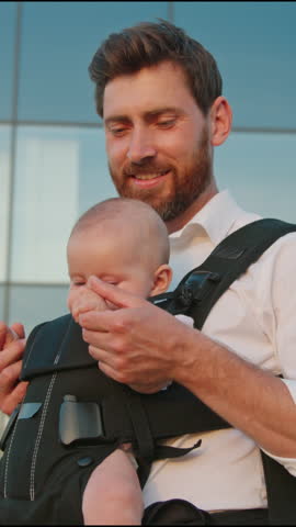 Cute portrait of funny newborn baby in a slings carrier. Young father holding a child kiss and hug on street. Ergo backpack. Parents. Slow motion