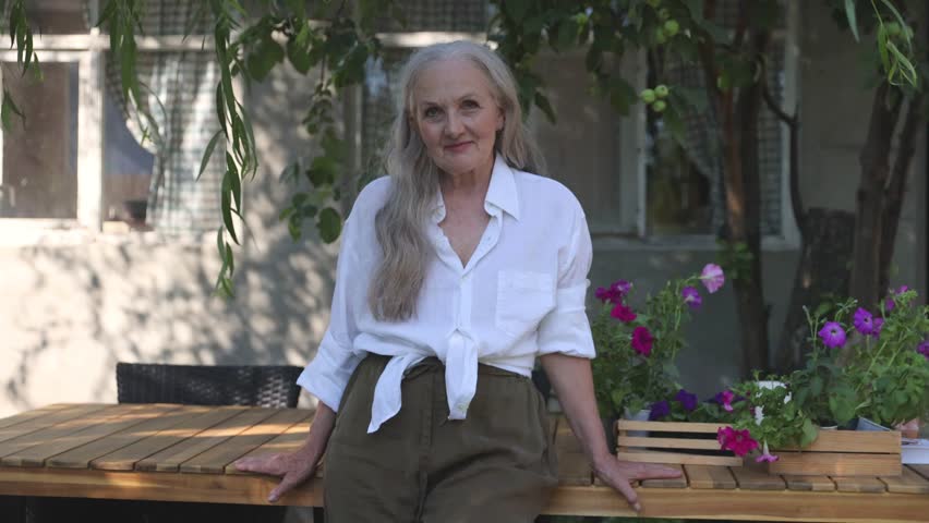  elderly woman  with long grey hair looks at the camera standing in the garden near the house. 