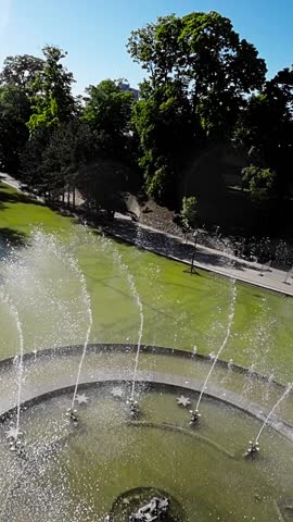 The video shows a fountain in Opole, Poland. The fountain is located in a park and is surrounded by trees and grass. The water in the fountain is green and there are several jets of water spraying