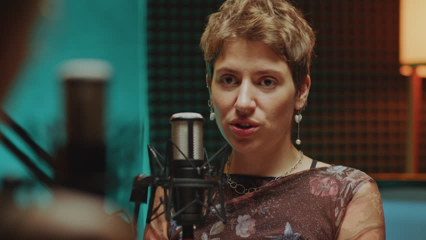 Female podcast guest sitting in recording studio and talking into microphone while being interviewed by host. Over-the-shoulder view