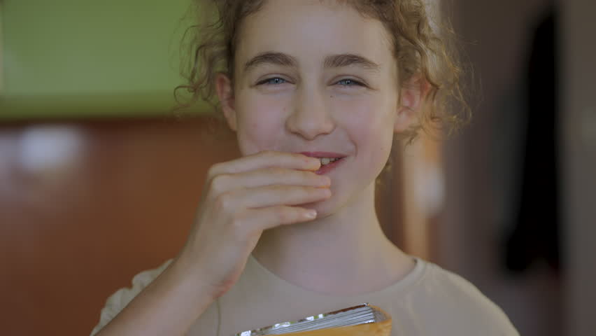 Hungry Girl Enjoying Tasty Chips. Kid Eating Unhealthy Potatoes Crisps. Snack on Junk Food. Portrait of Girl With Satisfied Face. Child Happily Eats Fast Food, Junk Food at Home. Childhood Obesity.
