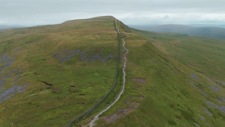 Drone view of a long path winding through green hills under a cloudy sky