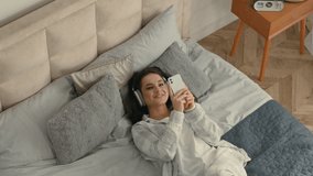 Top view young happy brunette woman lying on bed headphones listening music using smartphone mobile phone technology home inside comfort cozy joyful relaxed satisfied girl enjoy smile laugh pleasure - Powered by Shutterstock - Get 15% off with code: PIKWIZARD15