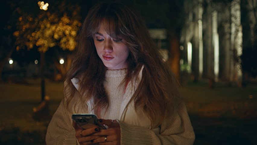 Walking girl texting smartphone at night city closeup. Serious woman looking cellphone screen strolling at illuminated by streetlights town. Pretty lady holding mobile phone alone in dark street.