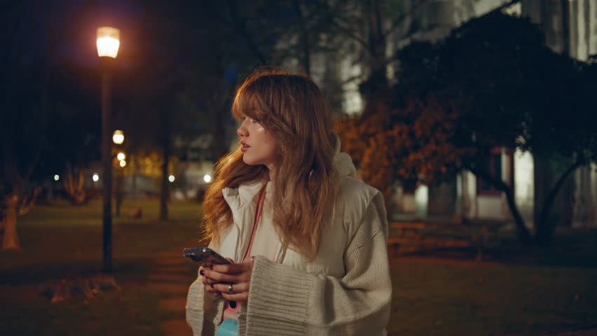Woman with smartphone standing night street closeup. Attractive lady browsing cellphone application waiting taxi late evening. Relaxed girl looking mobile phone communicating online at dark city.