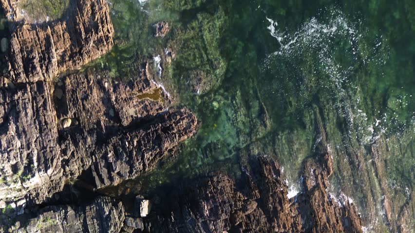 Aerial view in Monteferro, Nigrán showcasing rugged coastal rocks and clear turquoise waters. The scene begins near the water, gradually ascending to reveal the expansive rocky shoreline and ocean.