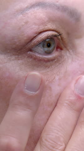 Senior woman massages undereye skin with gentle strokes on light background closeup. Grey haired old female enjoys rejuvenation skincare routine at home. Aged person everyday skin care procedure