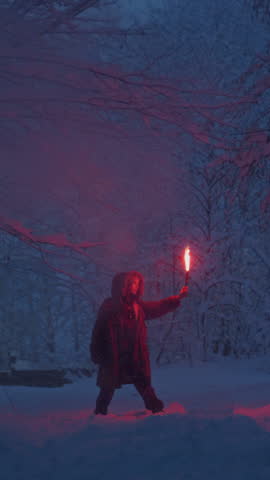 Portrait: A young girl holding a burning signal fire. A woman in a winter forest starts a red rescue flare. Snow on the trees, blue hour. Female person gives a distress signal, snowy weather