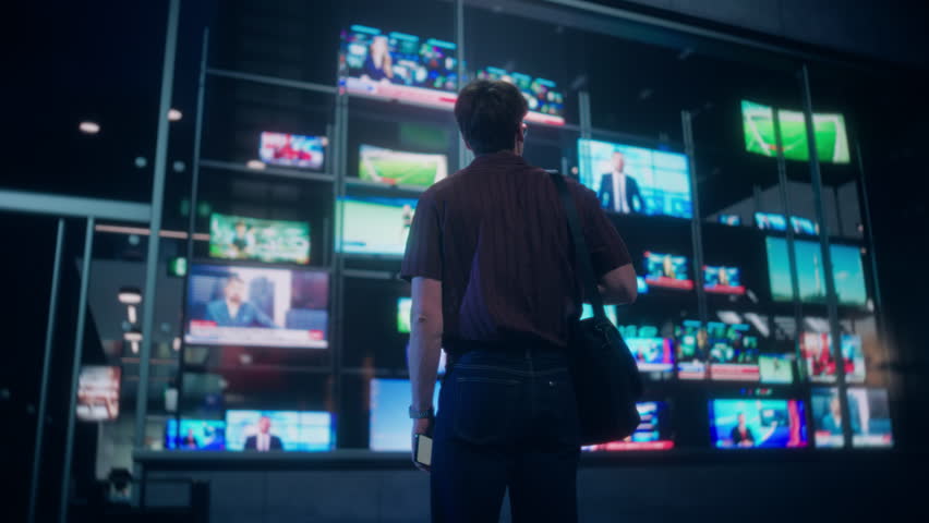 Young Caucasian Man with a Smartphone Stops to Look at a Vibrant Storefront TV Display with Diverse News and Entertainment Channels. Pedestrians Pass by on a Busy City Street at Night