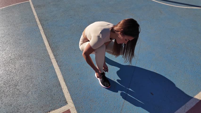 Sporty young woman tying running sneakers in urban park during summertime sunny day.