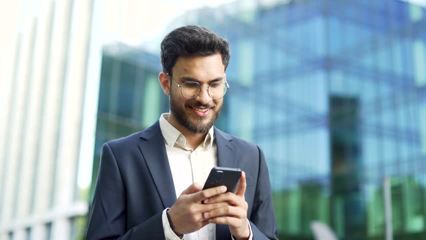 Confident businessman in suit stands near modern glass business office building holding smartphone in hands. Bearded man in glasses entrepreneur, executive or manager using a mobile phone outside