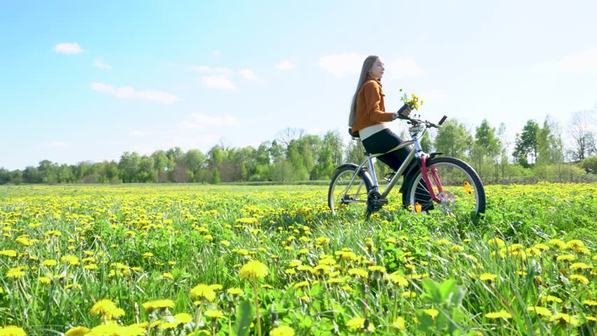 A young blonde girl sits on a bicycle among a dandelion field. She holds a bouquet and smells it. Enjoys spring and nature.