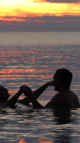 Happy marriage couple joyful hug and kissing in sea water at sunset time. View from back on sea waves background. Happy marriage and love concept. Valentine's Day in tropics