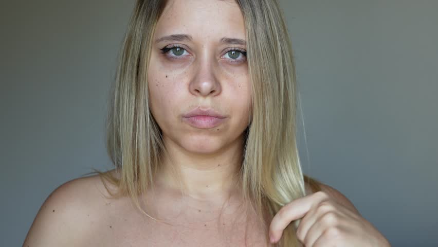 A young caucasian blonde woman with dry bleached hair holding scissors in her hands going to cut the ends of her hair on a grey background. Hair care