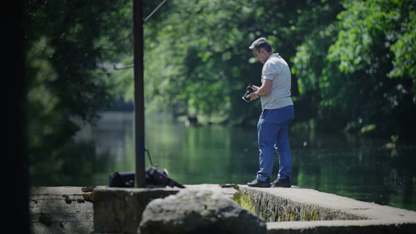 Photographer man is standing on a dock with camera looking at the water and taking a breathtaking landscape photos of the river enjoying the peaceful atmosphere