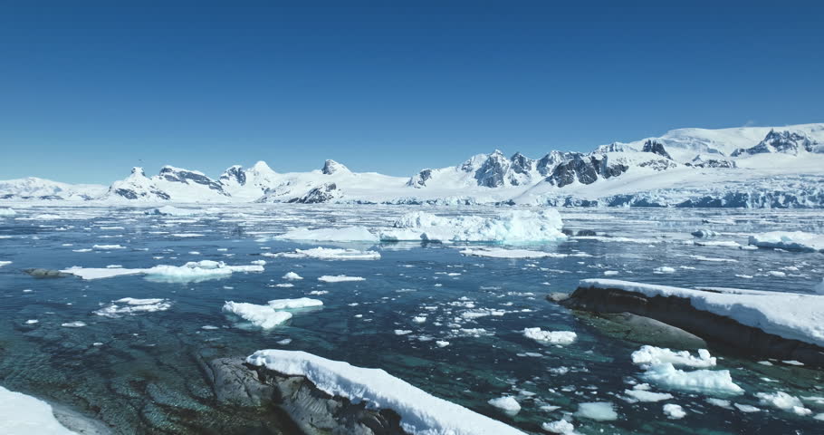 Fly over sunny Antarctic landscape, ocean rocky coast, melting icebergs, snow covered mountains in background. Arctic majestic wild nature seascape. Travel, explore Antarctica. Low angle drone flight