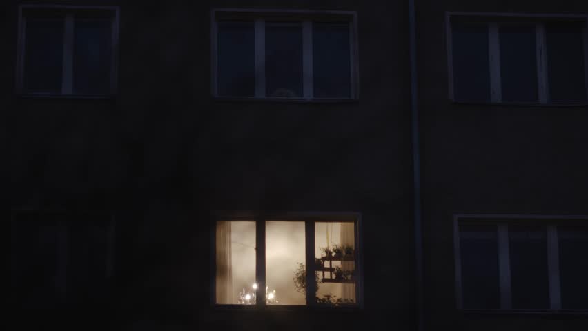 Evening view of a dimly lit apartment window glowing warmly, surrounded by darkness, creating a cozy and intimate ambiance.