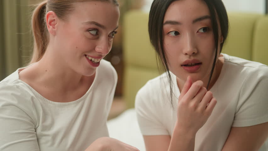 Multiracial friends women Caucasian woman whisper secret gossip talk secrecy information on ear to Asian Japanese female Chinese Korean girl laughing smiling talking confidential gossiping indoors