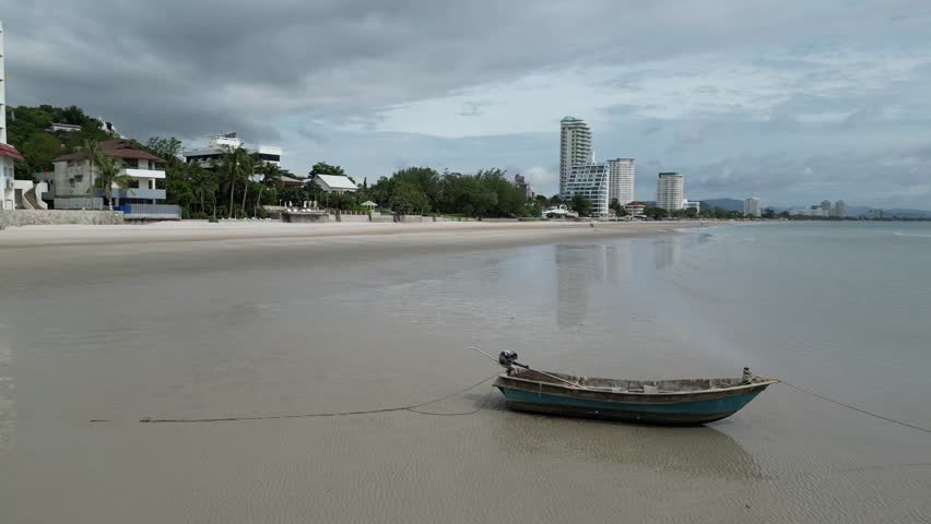 Small boat on wide sandy Hua Hin Beach near Chopstick Mountain. Skiff on shore at low tide in Thailand and beachfront condos and hotels. Aerial view from drone.