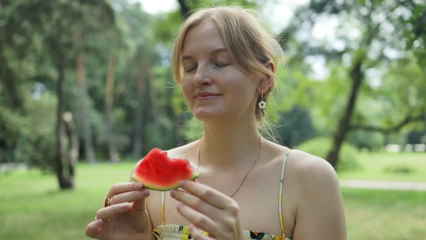 Charming blonde girl in summer dress eats and enjoys the taste of ripe red berry watermelon at a picnic in the city park. High quality FullHD footage