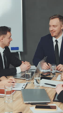 Vertical Screen: Business meeting with diverse team members shaking hands, discussing strategies, and collaborating at table with laptops and documents. Concept of business negotiation