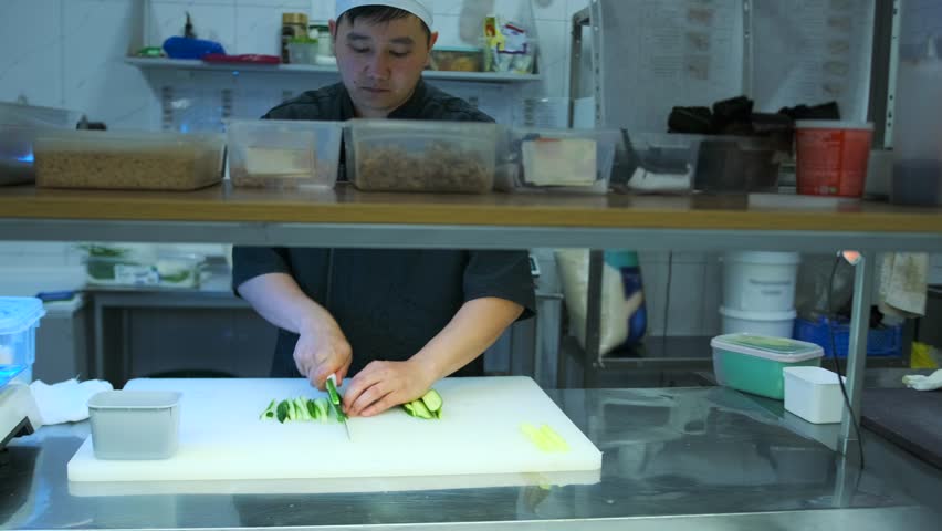 Asian man chef cutting fresh cucumber on slice for sushi rolls, on a cutting white board in a professional kitchen. Slow motion. Mid shot.