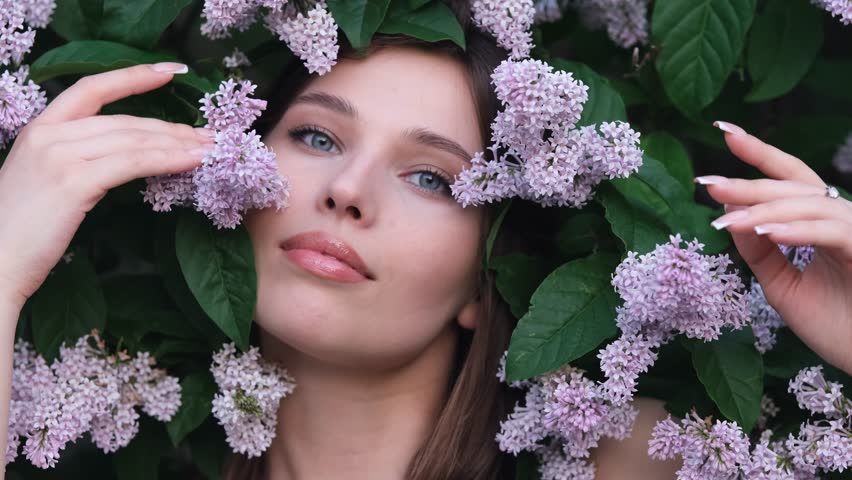 Close-up portrait of a girl in spring flowers. The face of a young woman is surrounded by lilac tassels. Beautiful girl among blooming lilacs.