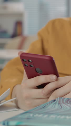 Vertical portrait of teenage Caucasian boy wearing yellow shirt sitting at desk in classroom using smartphone during lesson and cheerfully smiling