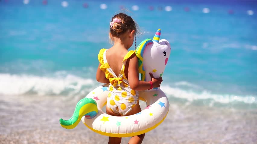 Summer vacation scene of a 5-year-old blonde girl walking on the beach with a swim ring around her waist, turquoise sea in the background. Capturing the joy and freedom of childhood during a sunny bea