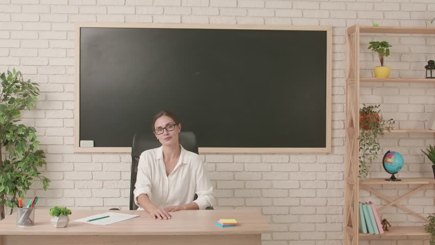 Woman college teacher sitting at desk in classroom in front of chalkboard pointing at empty area and shows thumb up. Education concept.