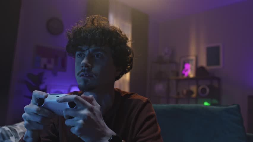 Young Caucasian gamer using game controller during videogame playthrough. Curly man enjoying spending time on digital entertainment. Using technology device while sitting on couch. Lifestyle concept.