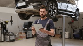Mechanic using digital tablet in car repair shop with raised vehicle in background. Man in work uniform checking diagnostics. Auto service, modern technology in automobile maintenance. - Powered by Shutterstock - Get 15% off with code: PIKWIZARD15