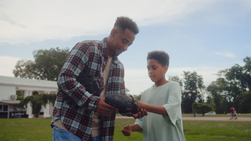 Careful father showing special gaming tools to son and getting ready for baseball game at family weekend at playground. Two generations of men preparing for training and using safety equipment.