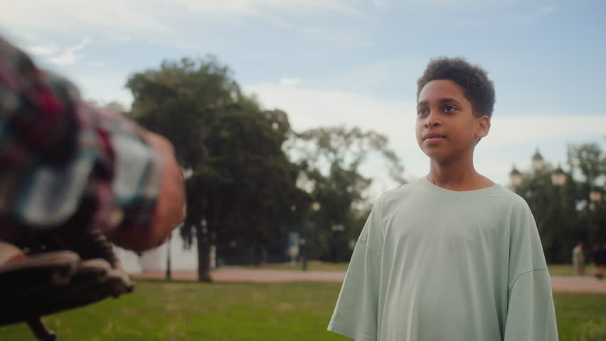 African american kid in oversize t-shirt giving high five to unrecognisable male in checkered shirt and smiling outdoors. Happy family members setting attitude and passing each other leather ball.