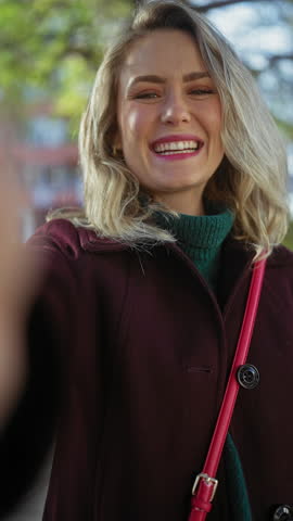 A young, happy caucasian woman with blonde hair, wearing a burgundy coat, posing for a lively outdoor portrait in a lush green city park.
