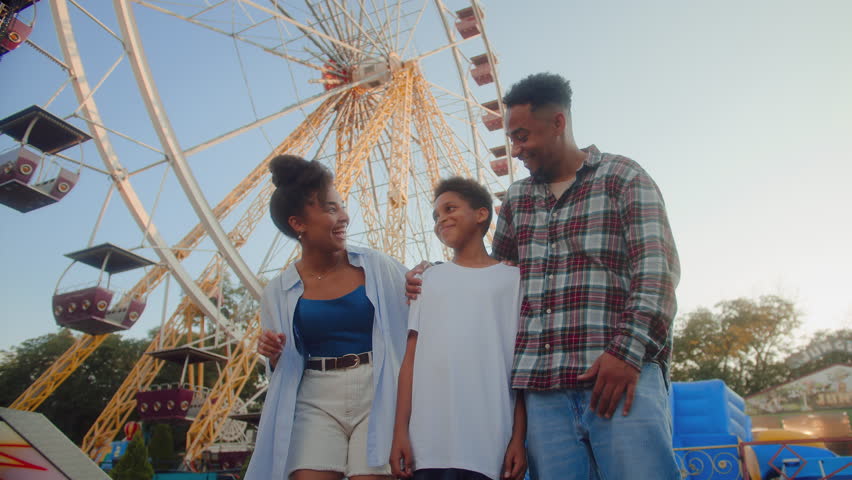 Two adults standing with smiling kid while looking aside at extreme roller coaster with excitement. African american family waiting for ride at popular attraction by carousel and ferris wheel.