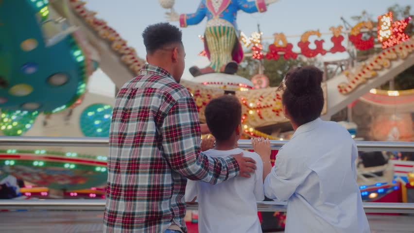 Back view family members wearing cotton shirt while watching kids attraction spinning and glowing at festive event outdoors. Playful parents and child spending good quality time together.
