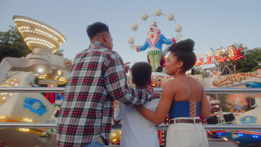 Back view of three people standing by fence of attraction with multicoloured flashlights and laughing emotionally. Excited people observing rotations of roller coaster and waiting for ride.