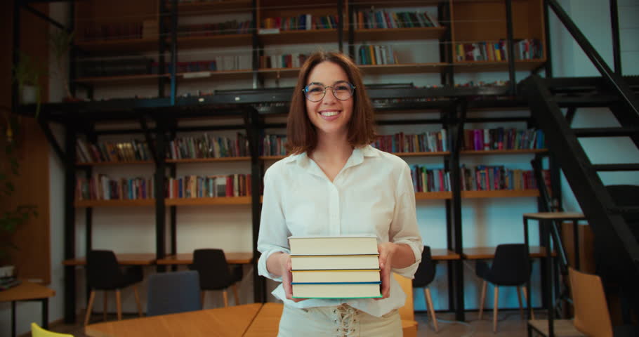 Portrait of a happy brunette girl in round glasses and a white shirt who walks with books in her hands through a library with modern books