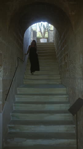A young woman descends a stone staircase inside a typical italian masseria, adorned with rustic walls and outdoor light streaming through the archway.