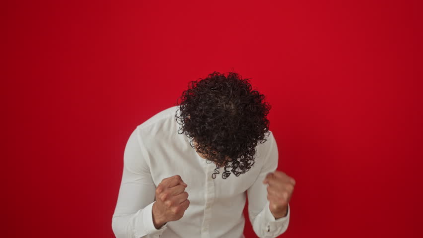 Young, excited hispanic man in shirt standing as a winner. success and achievement gesture with arms raised over isolated red background. vibrant, joyful scream celebrating win.