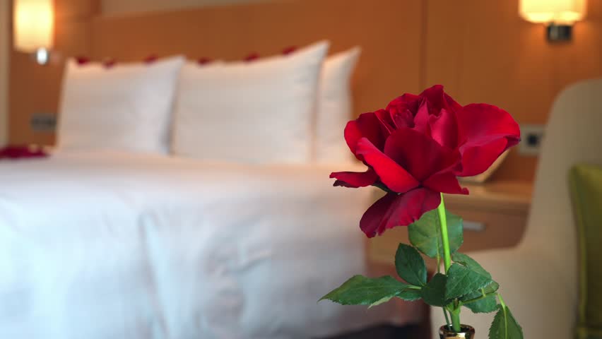 A bedroom with white bed and a table holding a gold vase with a single rose with empty copy space. The decor is romantic, ideal for a Valentine Day celebration or an anniversary surprise.