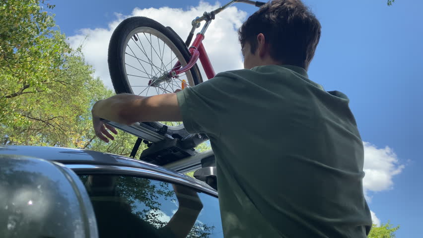 Teenage boy putting bicycle on car roof for trip. Car with roof bike rack transporting folding bike on the roof. Roof mount for carrying bikes.