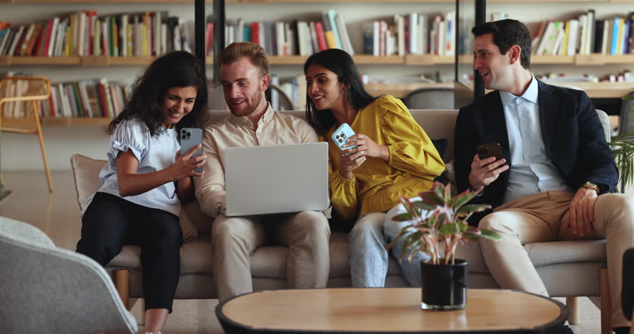 Cheerful diverse team of friends using gadgets in co-working space, showing online content to each others, discussing social media news, funny videos. Group of happy students enjoying break in library