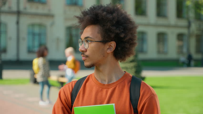 Serious calm African American student guy man male in eyeglasses holding book posing standing in city outdoors university college academy campus high school looking at camera education studying learn