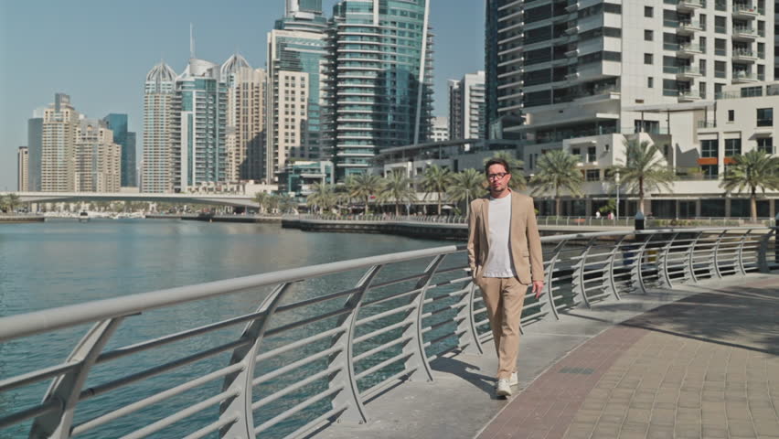 relaxed rich businessman walking on embankment in center of Dubai, admiring beautiful skyscrapers in modern business district in downtown, success and happy life of business owner