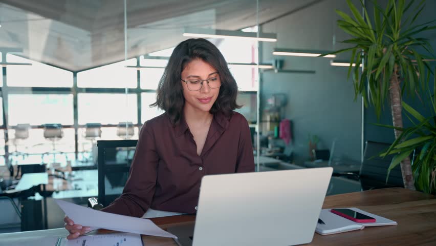 Accountant entrepreneur manager businesswoman doing paperwork using pc. Smiling latin hispanic young business woman working on laptop computer reading financial document report in office. Copy space