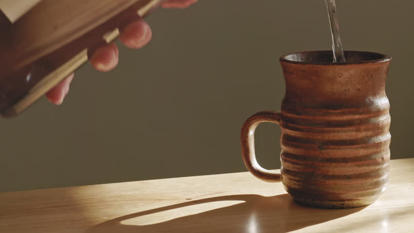 A hand pouring water from a transparent jug into a clay brown cup with sunlight